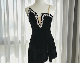 Könnte beinhalten: Ein schwarzes Samtkleid mit asymmetrischem Ausschnitt und tiefem V-förmigen Schlitz, verziert mit schillernden Kristallen an den Rändern und einem hautfarbenen Netzeinsatz. Das Kleid hat dünne beige Träger und einen mit kleinen Kristallen besetzten Saum.