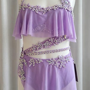 May include: A lavender two-piece dance costume featuring a halter neck top with ruffled chiffon layers and intricate floral lace appliqués adorned with clear crystals. The matching skirt has a side-swept chiffon overlay and crystal embellishments.