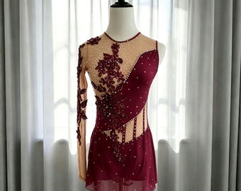 May include: A burgundy and nude leotard featuring a single long sleeve, intricate floral lace appliqués, and scattered clear crystals. The garment includes a sheer mesh bodice with a diagonal design and a layered skirt with a delicate finish.