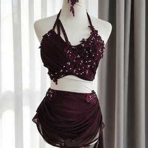May include: A deep burgundy two-piece dance costume featuring a halter-style crop top with intricate floral lace appliques, shimmering sequins, and beads. The matching skirt has a draped, layered chiffon overlay with a side slit and sequin trim.
