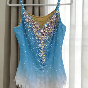 May include: A light blue leotard with a white ombre hem and a jagged edge. The bodice features a nude mesh neckline and is heavily embellished with iridescent crystals and larger faceted stones arranged in a central vertical pattern.