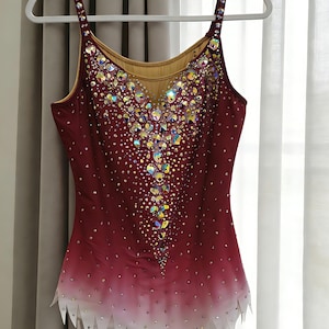 May include: A sleeveless leotard featuring a deep burgundy to white ombre gradient. The bodice is heavily embellished with iridescent crystals in a central vertical pattern, with smaller scattered crystals throughout and a jagged white hemline.