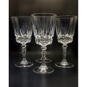 May include: Four clear crystal wine glasses with a cut glass design. The glasses have a faceted bowl, a slender stem, and a round base. The glasses are arranged on a dark surface, and the light reflects off the crystal.