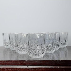 May include: A set of eight clear, faceted drinking glasses. The glasses are arranged in a row on a dark wooden surface. The glasses have a diamond pattern on the lower half and vertical lines on the upper half.
