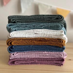 May include: A stack of seven folded, textured fabric items in various colors. The colors include dark green, teal, white, mustard yellow, navy blue, light pink, and deep purple. The items are stacked on top of each other, with the edges of each item visible.
