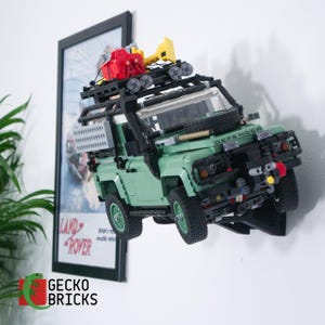 May include: A sage green Land Rover Defender model vehicle constructed from plastic building bricks, mounted on a black wall bracket. The model features a black roof rack with a red storage box, yellow accessories, and a spare tyre on the bonnet.