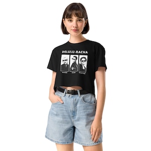 May include: Black crop top featuring the text "DELULU-RACHA" above three black and white portraits. The portraits are labelled "THE FATHER", "THE SON", and "THE HOLY TOAST". The model is wearing denim shorts.