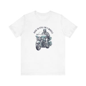 Puede incluir: Camiseta blanca con un gráfico de motocicleta y un piloto. El texto "OVER 70 STILL ON 2 WHEELS" está encima de la motocicleta en un diseño circular. La motocicleta es de color azul claro. Un regalo para moteros.