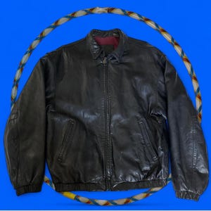 Pode incluir: Casaco de couro preto com fecho de correr, dois bolsos frontais e bainha e punhos canelados. O casaco tem gola e forro bordô. Um bambolê colorido envolve o casaco sobre um fundo azul.
