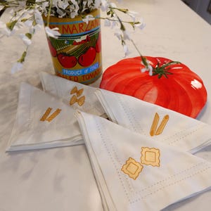 Puede incluir: Naturaleza muerta con servilletas de tela blanca bordadas con formas de pasta, un plato con forma de tomate y una lata de tomates italianos. Las servilletas tienen detalles bordados de pasta de lazo, penne y ravioles.