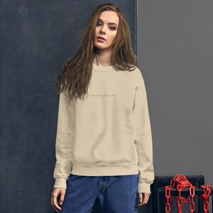 Op de afbeelding: Een beige sweatshirt met de tekst "currently processing" in een lichtbruin lettertype. De sweatshirt heeft een ronde hals en is gemaakt van een zacht materiaal. Het model draagt een blauwe jeans.