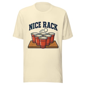 Puede incluir: Camiseta color crema con un diseño en azul marino que muestra vasos rojos y una pelota de ping pong, con el texto "NICE RACK". El diseño está centrado en la parte delantera de la camiseta.