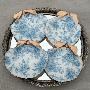 Puede incluir: Cuatro posavasos en forma de concha con un estampado floral blanco y azul, dispuestos sobre una bandeja de espejo plateada. Los bordes de los posavasos están adornados con un color dorado metálico.