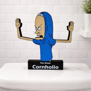 May include: A novelty toilet paper holder featuring a cartoon character in a blue robe with arms outstretched. The base is black with the words "The Great Cornholio" in white. The holder is placed on a white toilet.