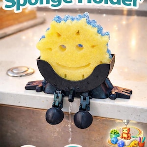 May include: A yellow sponge with a smiling face rests in a black sponge holder. The holder has legs and a curved design, with the words "Cute Sponge Holder" above. The image also includes icons for anti-rust and fast drying.