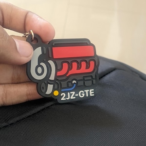 Puede incluir: Un llavero negro, rojo y gris con un diseño de motor estilizado. El llavero tiene el texto "2JZ-GTE" en blanco. El llavero está sujeto a un anillo de metal.