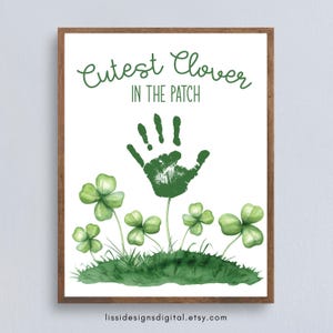 May include: A framed print with a white background features green watercolor clovers and grass. The text "Cutest Clover IN THE PATCH" is above a green handprint. The Etsy shop URL is at the bottom.