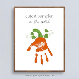 May include: A framed wall art print featuring the text "cutest pumpkin in the patch" in black lettering above an orange handprint shaped like a pumpkin with a green stem and vine accents. The design is centered on a white background.
