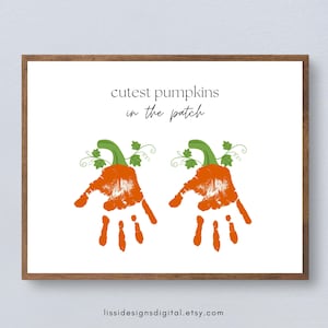 Può includere: Una stampa rettangolare bianca con il testo nero "cutest pumpkins in the patch" sopra due illustrazioni di impronte di mani arancioni. Ogni impronta è sormontata da un picciolo di zucca verde e viticci, incorniciata da un bordo in legno scuro.
