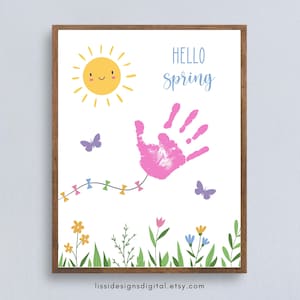 Könnte beinhalten: Ein gerahmter Druck mit Frühlingsmotiv. Er zeigt eine lächelnde gelbe Sonne, einen rosa Handabdruck und die Worte "Hello Spring" in Blau. Schmetterlinge und eine Girlande aus bunten Fähnchen sind ebenfalls enthalten, zusammen mit einem floralen Rand.
