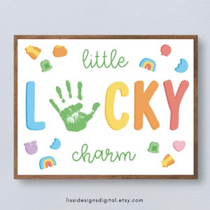 Pode incluir: Uma impressão emoldurada com as palavras "little LUCKY charm" em letras coloridas e uma impressão de mão verde. A impressão é decorada com vários amuletos da sorte, incluindo trevos, arco-íris e estrelas, em um fundo branco.