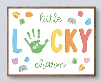 Può includere: Stampa incorniciata con le parole "little LUCKY charm" in lettere colorate e un'impronta di mano verde. La stampa è decorata con vari portafortuna, tra cui quadrifogli, arcobaleni e stelle, su sfondo bianco.