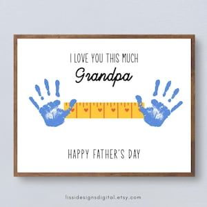 Peut inclure: Une impression rectangulaire dans un cadre en bois avec le texte I LOVE YOU THIS MUCH Grandpa et HAPPY FATHER'S DAY. Deux empreintes de mains bleues sont placées de chaque côté d'un graphique de règle jaune orné de petits cœurs rouges.