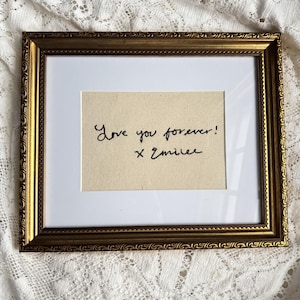 May include: A rectangular picture frame with an ornate gold finish and a white mat. Inside the frame, a piece of cream-colored fabric features black embroidered cursive text that reads, "Love you forever! x Emilee."