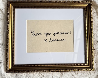 May include: A rectangular picture frame with an ornate gold finish and a white mat. Inside the frame, a piece of cream-colored fabric features black embroidered cursive text that reads, "Love you forever! x Emilee."