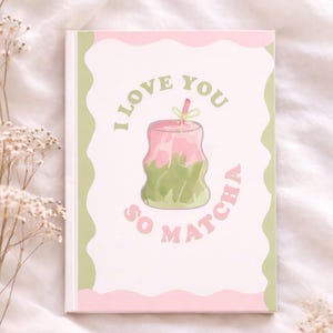 May include: A notebook with a white cover featuring the text "I LOVE YOU SO MATCHA" in pink and green. The design includes a pink and green matcha drink illustration. The notebook has a pink border and green wavy accents.