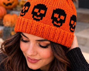 May include: A bright orange knit beanie featuring a repeating pattern of black skulls around the crown. The hat has a thick ribbed cuff and is topped with a large, fluffy, multi-toned orange and black faux fur pom-pom on the very top.