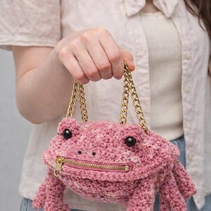 May include: A pink crochet frog-shaped handbag with a gold zip and chain straps. The bag has black eyes and frog feet. A unique accessory. The bag is a unique accessory.