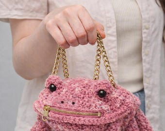 May include: A pink crochet frog-shaped handbag with a gold zipper and chain straps. The bag features black eyes and detailed frog feet. The bag is a unique accessory.