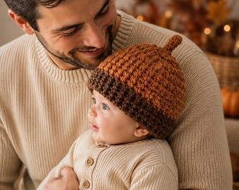May include: A crochet baby hat shaped like an acorn, featuring a textured, light brown upper section and a dark brown ribbed brim. The hat has a small, pointed stem at the very top, mimicking the appearance of a natural acorn cap and nut.