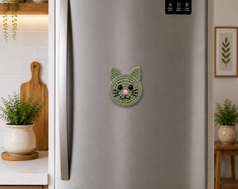 May include: A crocheted cat face magnet in shades of green, with a pink nose and black whiskers, is affixed to a stainless steel refrigerator door. The handmade magnet is round and features a smiling cat face.
