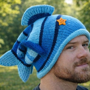 Könnte beinhalten: Eine blaue Strickmütze in Form eines Fisches, mit Flossen und einem Schwanz. Die Mütze hat verschiedene Blautöne mit dunkelblauen Streifen und einem kleinen orangefarbenen Stern. Die Mütze wird von einer Person getragen.