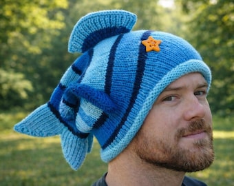 Puede incluir: Un gorro azul tejido en forma de pez, con aletas y cola. El gorro tiene diferentes tonos de azul con rayas azul oscuro y una pequeña estrella naranja. El gorro lo lleva una persona.