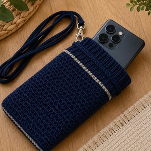 Könnte beinhalten: Marineblau gestrickte Handyhülle mit silbernem Streifen. Die Hülle hat ein passendes marineblaues Schlüsselband und einen silbernen Verschluss. Ein schwarzes Smartphone ist teilweise in der Hülle sichtbar. Die Hülle liegt auf einer Holzoberfläche.