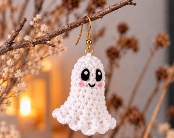 May include: A white crocheted ghost earring featuring a gold-toned metal hook. The ghost has two large black oval eyes, a small black stitched smile, and two small pink circular cheeks embroidered onto the textured white yarn surface.