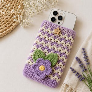 Puede incluir: Una funda de ganchillo para tel&eacute;fono con un patr&oacute;n de chevr&oacute;n en color lavanda y crema. La funda est&aacute; decorada con una gran flor de ganchillo morada con centro amarillo y dos hojas verdes. Un peque&ntilde;o bot&oacute;n de madera en forma de estrella cierra.