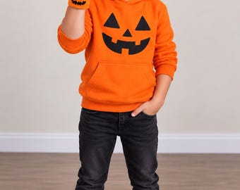 May include: An orange hooded sweatshirt featuring a black jack-o'-lantern face graphic on the chest and a front kangaroo pocket, paired with black denim jeans, white sneakers, and an orange crocheted wristband with a black jack-o'-lantern face.
