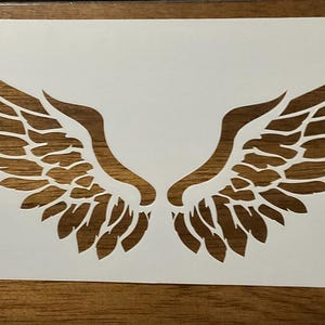 May include: A white rectangular stencil featuring a symmetrical pair of detailed, feathered wings cut out from the center. The design displays intricate layered feather shapes that curve upwards and outwards, creating a clear negative space pattern.
