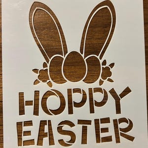 May include: White stencil featuring a bunny ears design with three egg shapes and floral accents. The words "HOPPY EASTER" are cut out below the design, perfect for Easter crafts and decorations.
