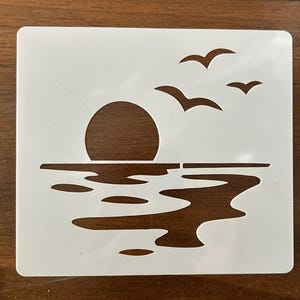 May include: A white plastic stencil featuring a sunset scene. The design includes a circular sun setting over a body of water, with three birds flying above. The water is depicted with wavy lines, creating a tranquil, nature-themed design.