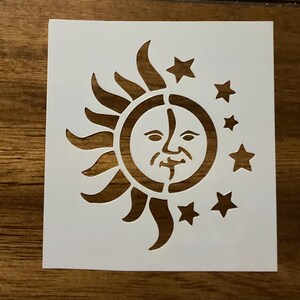 May include: A square white stencil featuring a cut-out design of a sun with a human face on the left side, surrounded by curved rays, and a cluster of five five-pointed stars of varying sizes arranged on the right side of the sun.