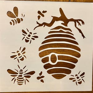 May include: A white stencil featuring a beehive and several bees. The beehive is oval-shaped with horizontal lines, and the bees are depicted in various sizes and poses. The stencil is designed for crafting and decorative purposes.