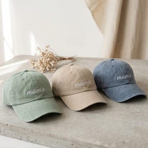 May include: Three baseball caps in sage green, tan, and blue-grey. Each cap has the word "mama" embroidered in white script across the front. The caps are displayed on a concrete surface with dried flowers in the background.
