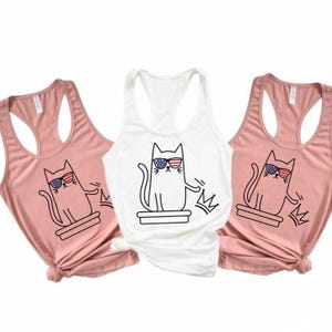 May include: Three tank tops: two in a dusty rose color and one in white. Each tank top features a black and white graphic of a cat wearing sunglasses with an American flag design. The cat is also wearing a crown.