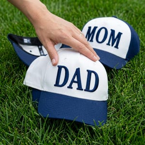 May include: White and navy blue baseball caps with the words "DAD" and "MOM" embroidered on the front. The caps are sitting on green grass.