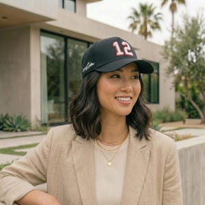 May include: Black baseball cap with the number 12 in red and white on the front. The word "custom" is written in cursive on the side. The cap is worn by a person wearing a beige blazer.
